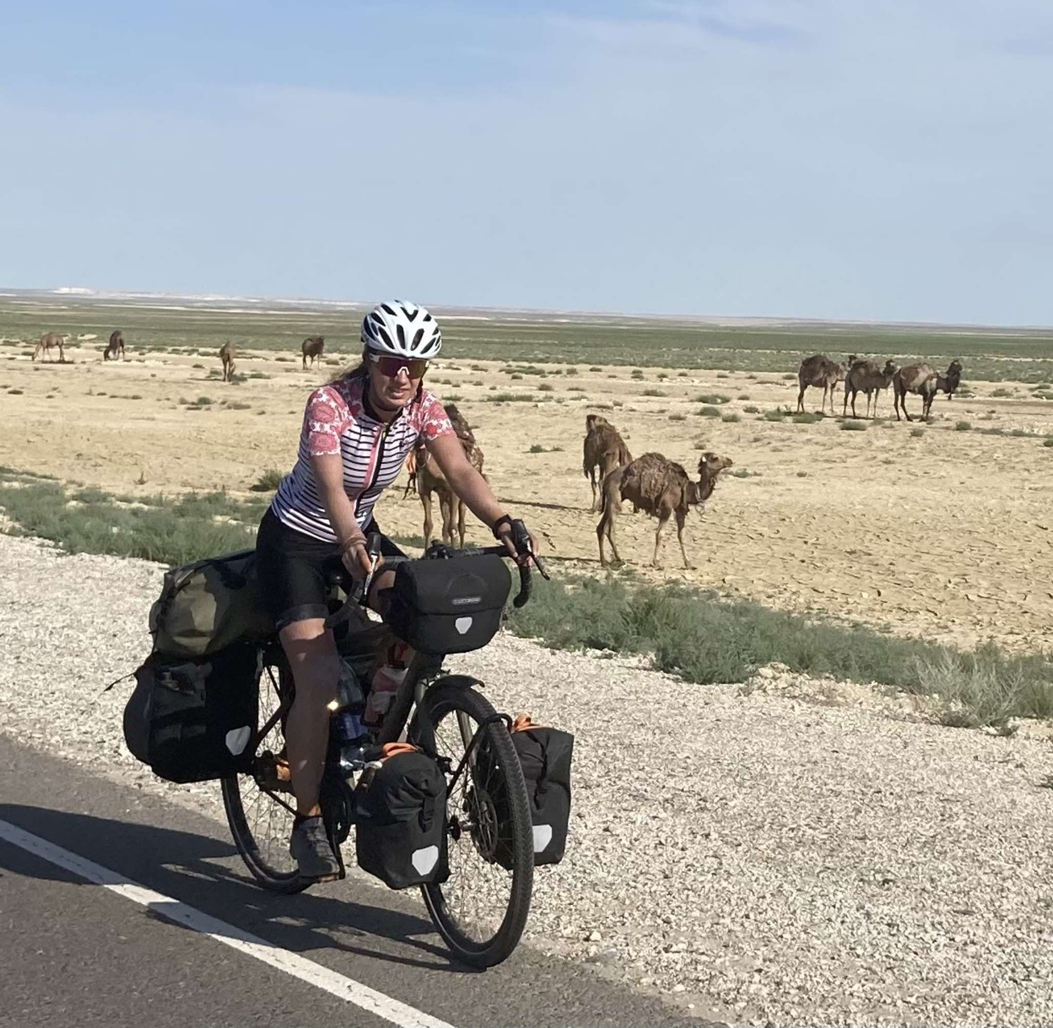 Marleen met volle bepakking fietst over een weg door een droge steppe met kamelen op de achtergrond.