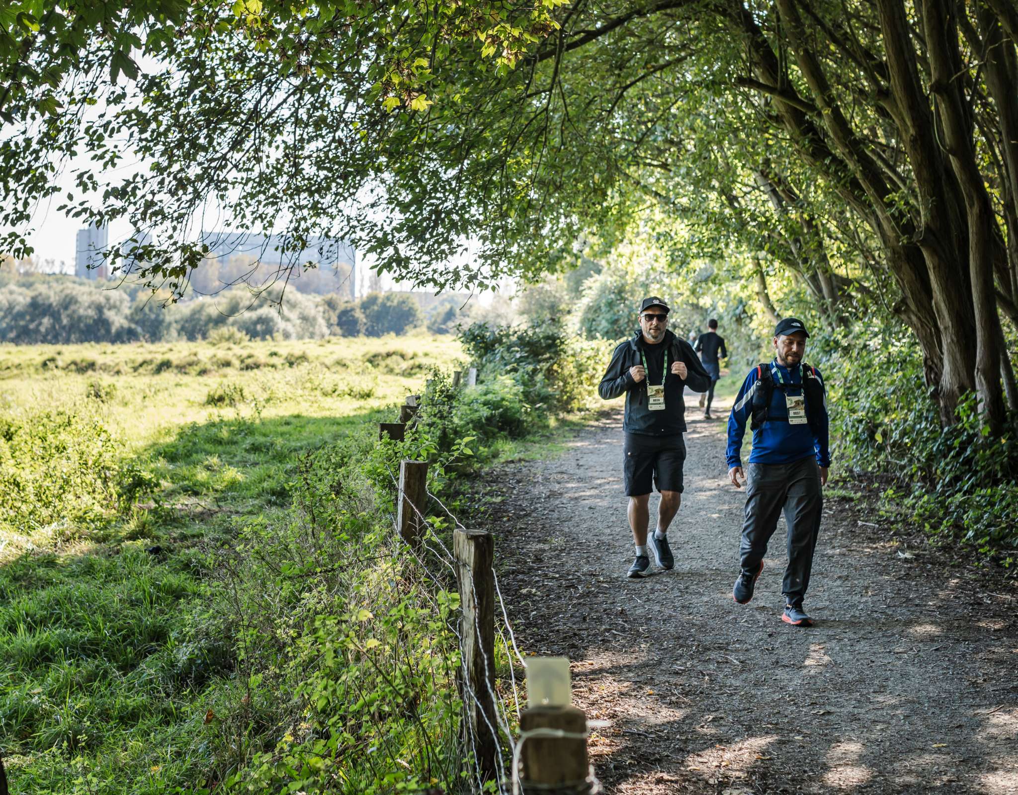 Twee wandelaars volgen een schaduwrijk pad langs groene velden.